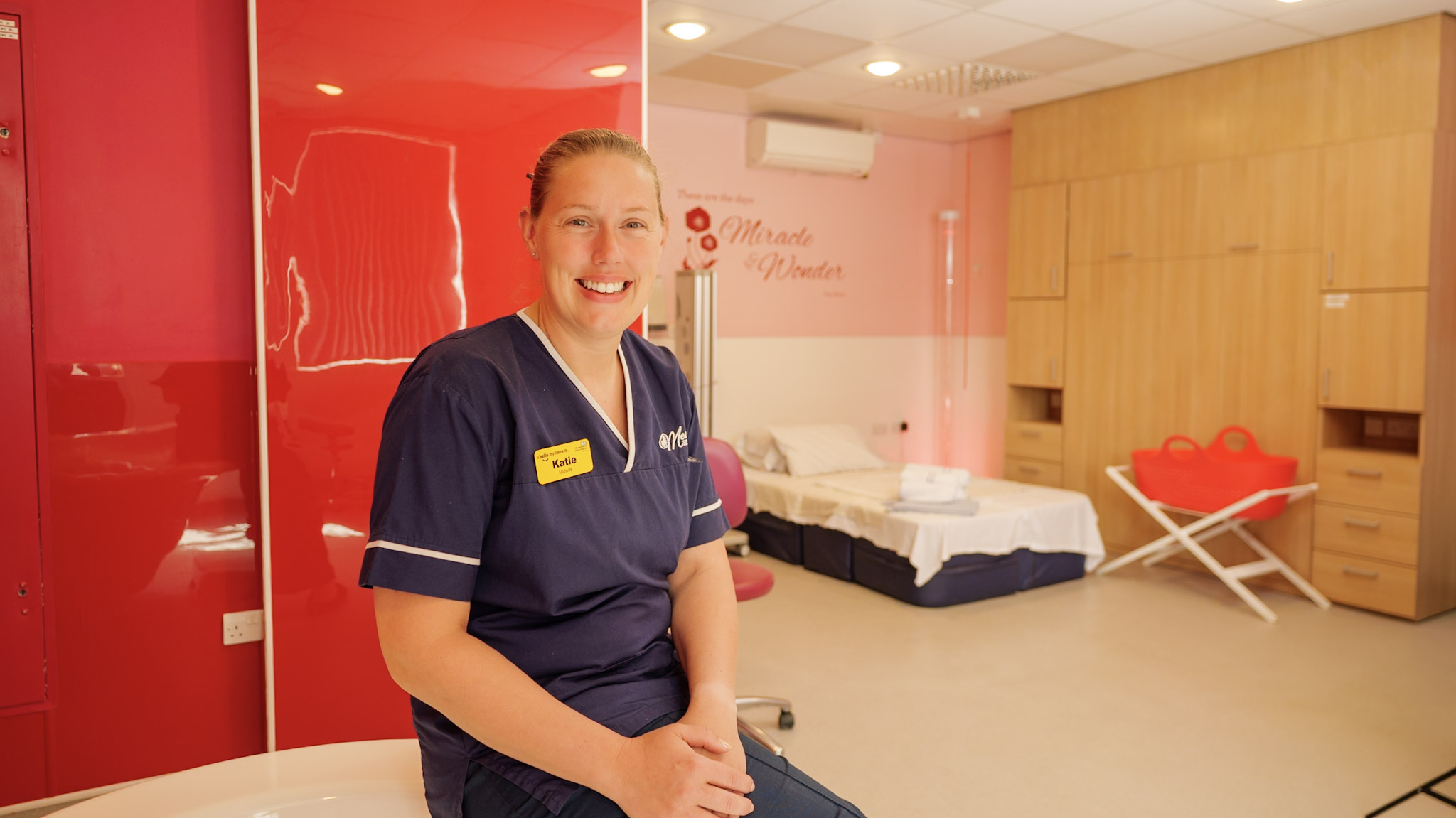 A midwife sat on the edge of a bathtub in a birthing suite with a bed behind them.