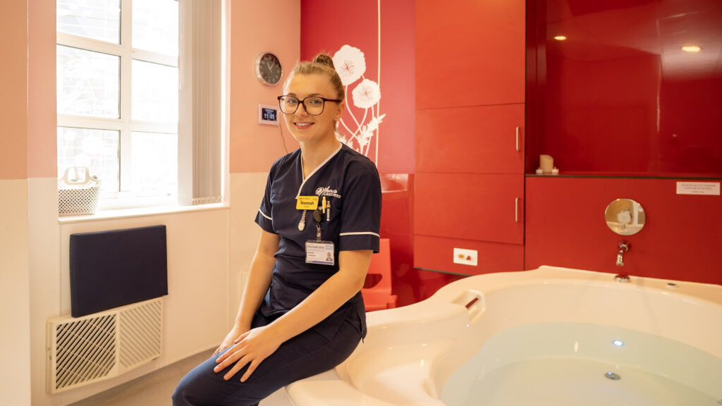 A midwife sat on the edge of a bathtub in a birthing suite.