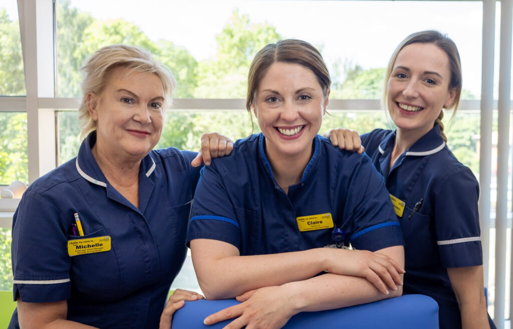 Three midwives in uniform stood smiling at the camera in a hospital setting