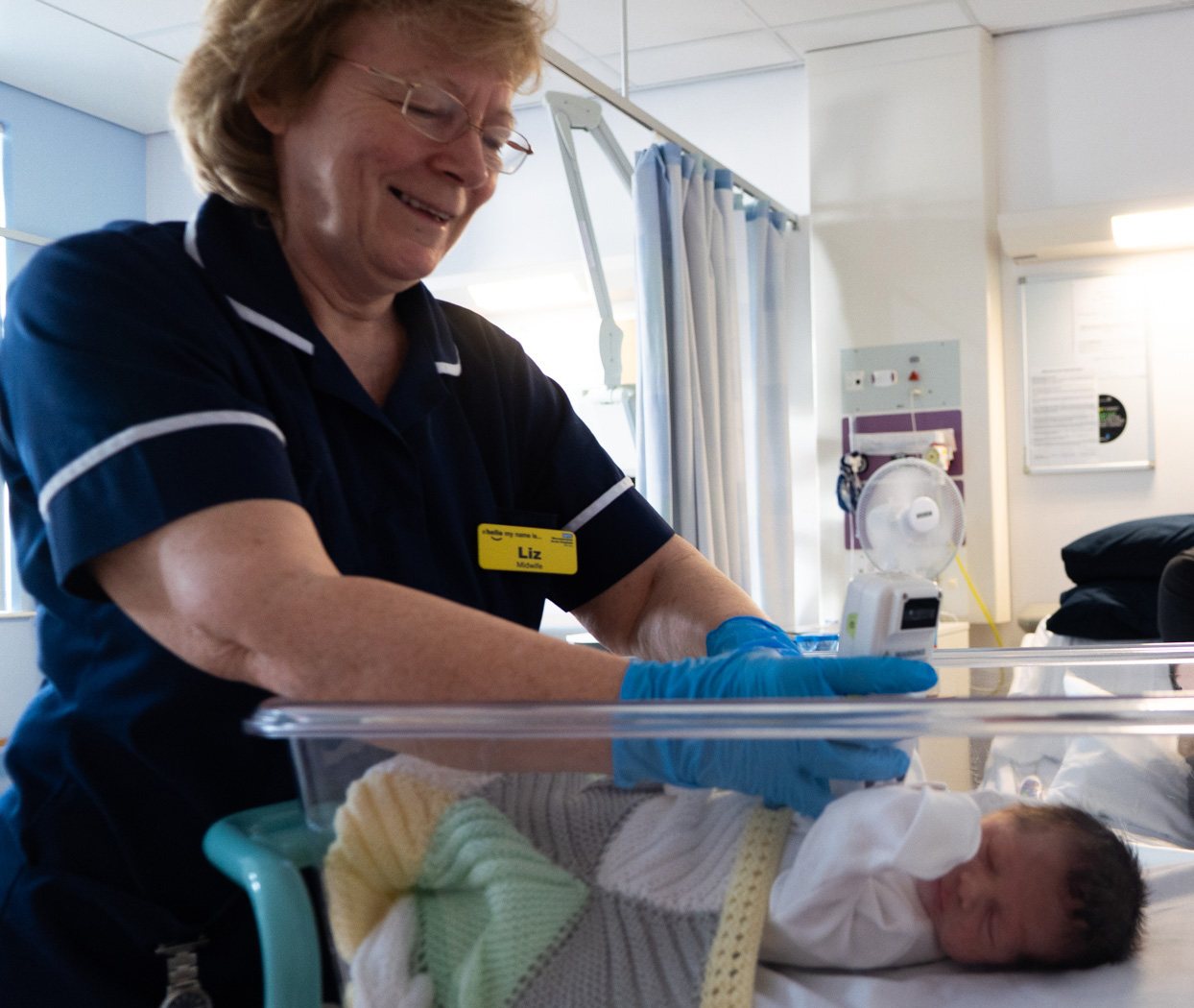 Midwife gently looking down at a sleeping newborn baby in a bassinet, with a maternity ward in the background.
