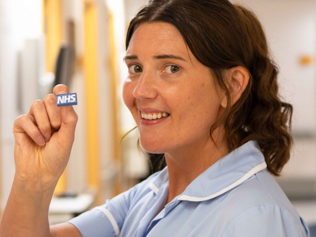 Portrait photo of nurse on hospital ward. Smiling holding an NHS badge.
