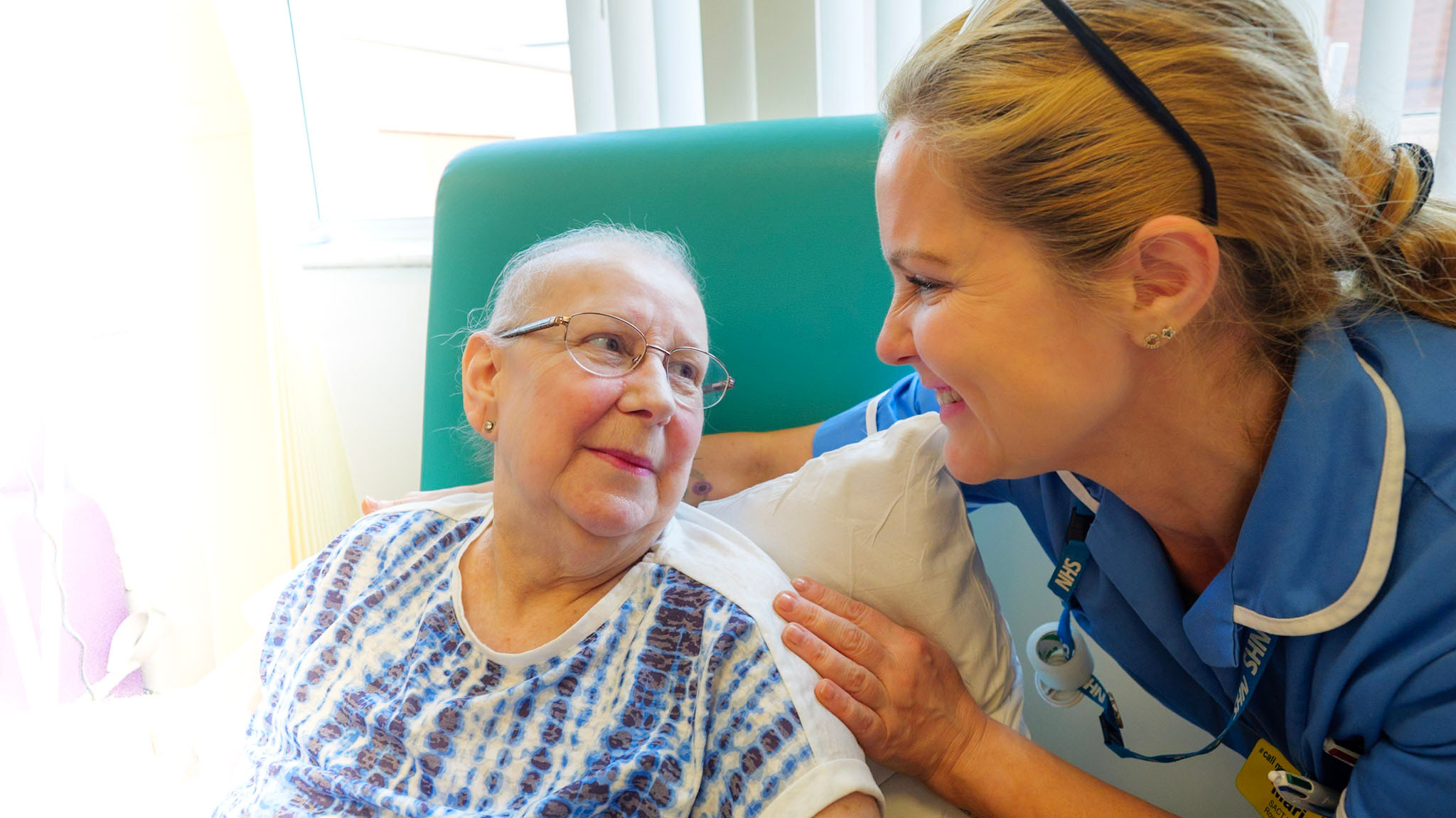 Photograph of nurse smiling at patient who is sitting in a chair. Both looking at one another and smiling.