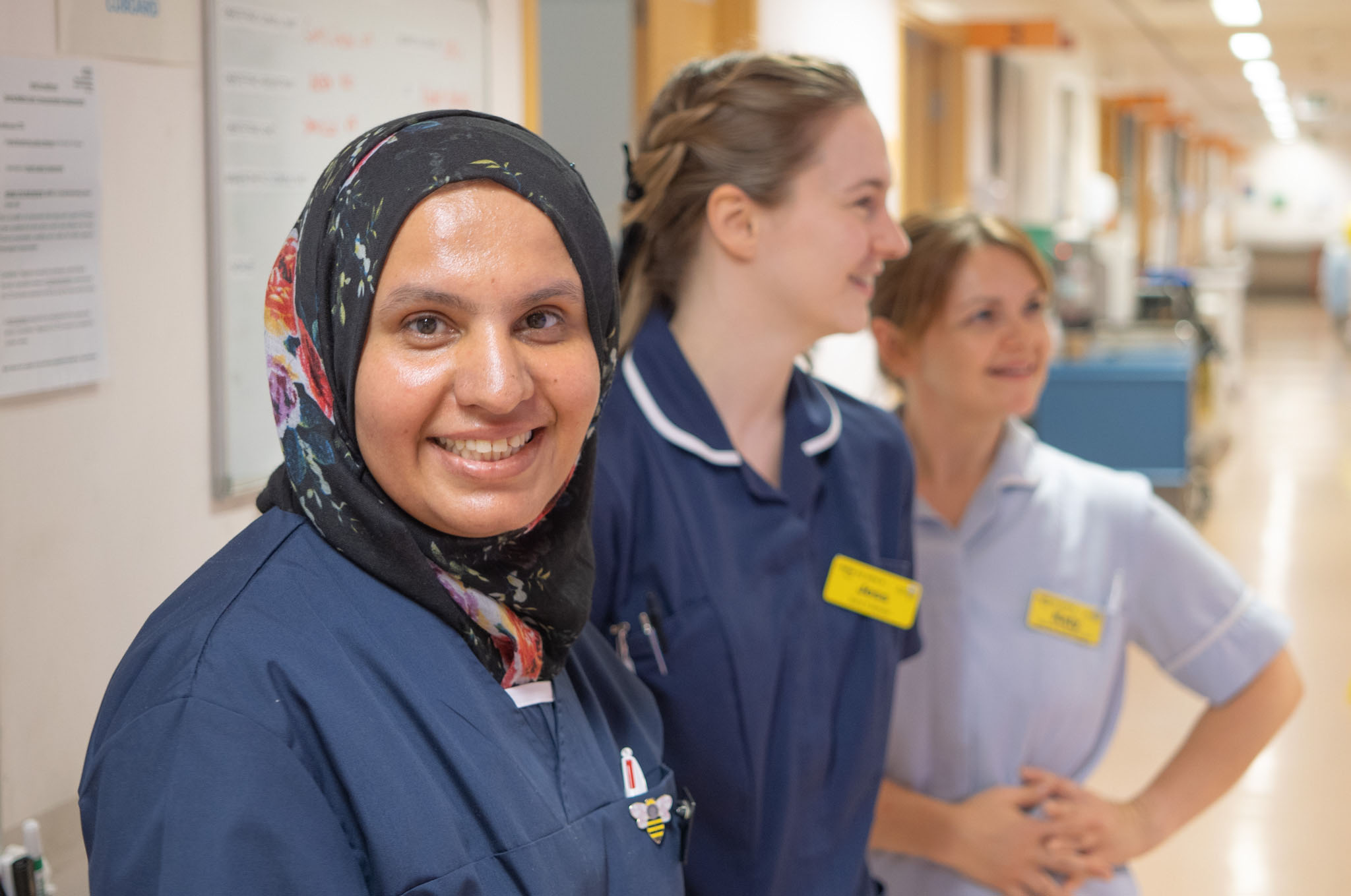 Two nurses and a support worker smiling together in a hospital setting, looking at something off-camera.