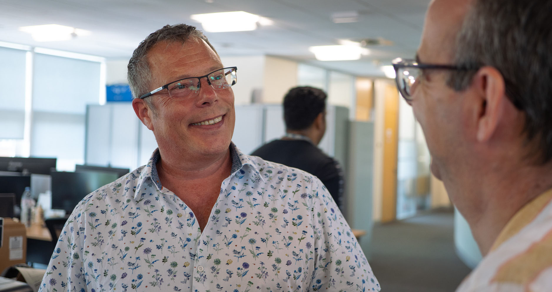 Photograph of two men talking to one another, smiling. Photograph taken in office with computer monitors behind.