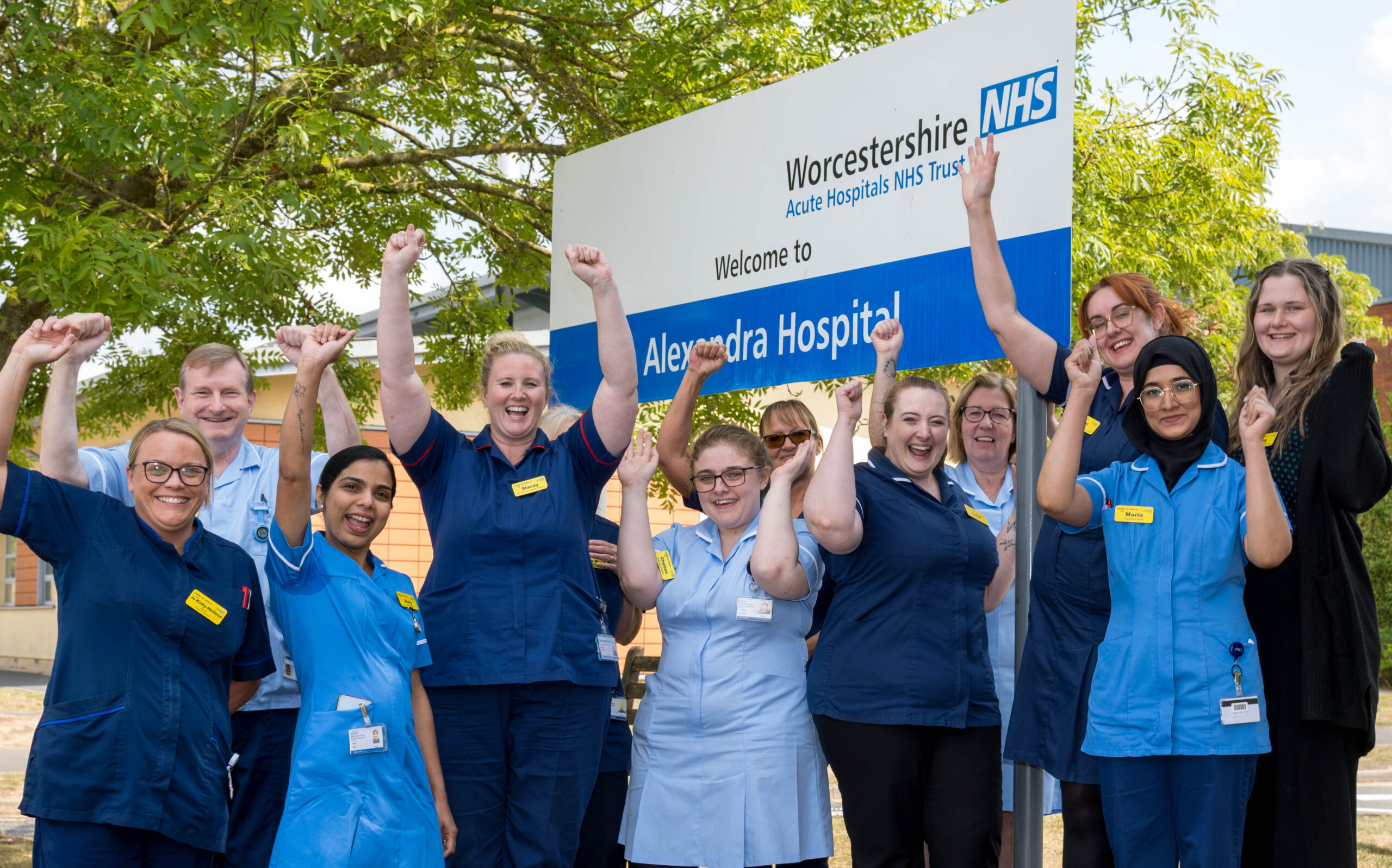 Hospital staff cheer with hands above their heads next to a sign for Alexandra Hospital Redditch