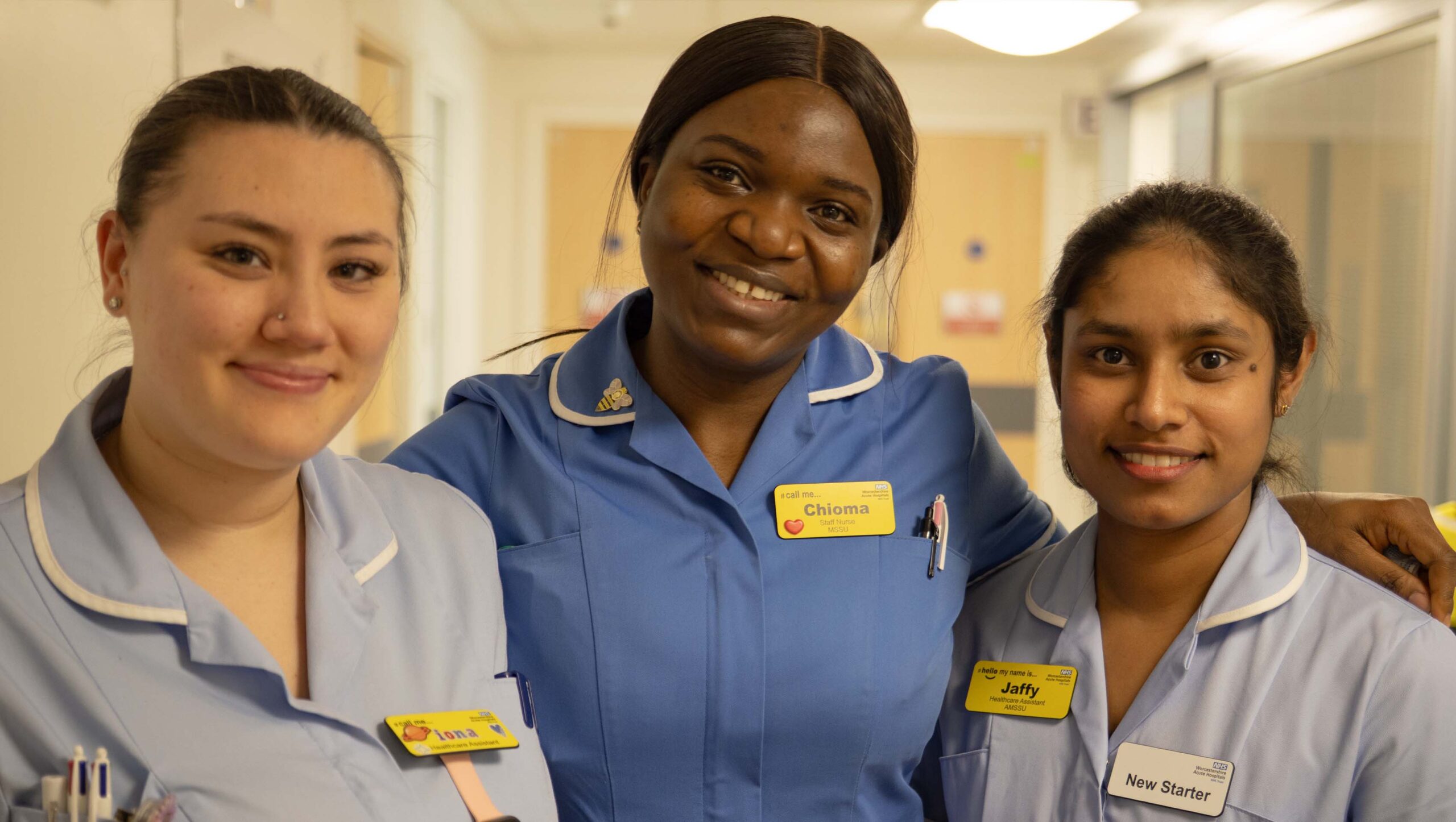 Close-up portrait of three hospital staff standing in a corridor and smiling at the camera. The central figure is a staff nurse, flanked by two Healthcare Assistants, one of whom is wearing a new starter badge.