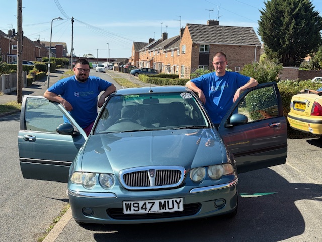Nick and Alex stood either side of Roger the car, which is a grey Rover parked on a residential street.