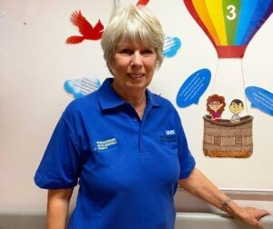 A woman in blue volunteer uniform stands in a children's hospital department