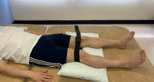 Image shows individual laying down flat with a belt looped and fastened just above the knees. Feet are shoulder width apart