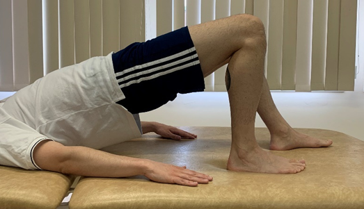 Image shows an individual in bridge pose with feet upper body, head and arms flat on floor and hips pushed up towards the ceiling. Affected leg (foot) is closer to body and other foot is slightly forward