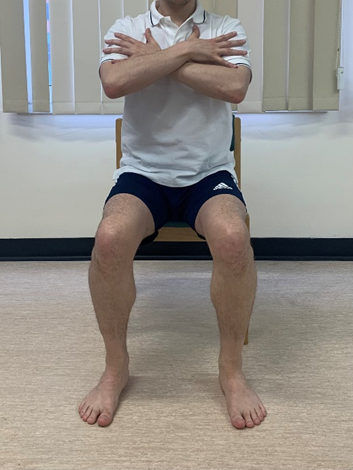 Image shows an individual sat on a dining chair feet flat on ground and arms crossed across chest