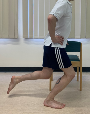Image shows an individual holding the back of a dining chair hand is on hip and one leg is raised from floor at a 90 degree angle. The other knee is slightly squatting