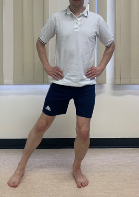Image shows an individual stood with hands on hips and feet waist width apart. One leg is placed straight out to the side with foot flat on floor. Other knee is bent to the front.