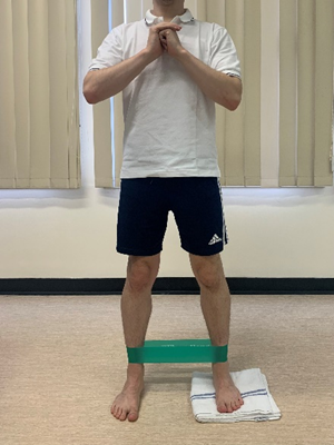 Image shows an individual stood on a hard floor with hands clasped at chest height to the front. One foot is on a folded towel and legs have a resistance band around them at just above ankle height. Legs are waist width apart