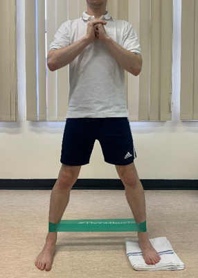 Image shows an individual stood on a hard floor with hands clasped at chest height to the front. One foot is on a folded towel and legs have a resistance band around them at just above ankle height. Legs are wider than waist width apart