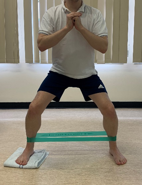 Image shows an individual stood on a hard floor with hands clasped at chest height to the front. One foot is on a folded towel and legs have a resistance band around them at just above ankle height. Legs are much wider than waist width apart and individual is in a squat stance 