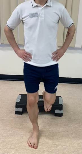 Image shows an individual stood on a hard floor with hands on hips. Feet a waist with apart and ball of one foot is resting behind on a raised step. Front leg is in a slight squat. They are stepping down from the step
