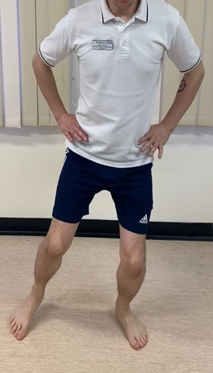 Image shows an individual stood on a hard floor with hands on hips. Feet a waist with apart and heel of one foot is raised and being stepped out to the side