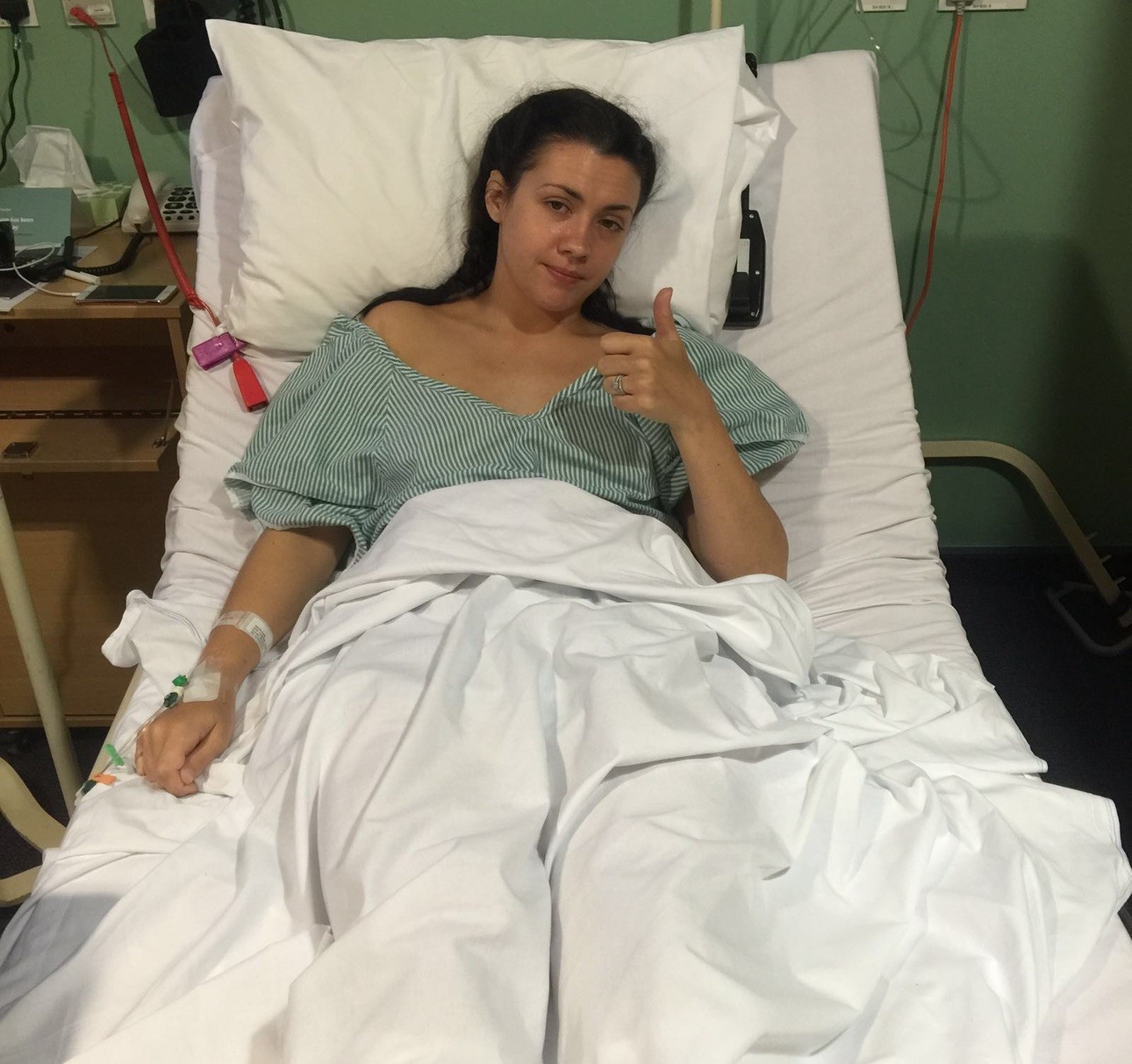 A woman lies in a hospital bed. Her legs are covered in a blanket. She has a cannula in one arm. She shows a thumbs up on her other hand.