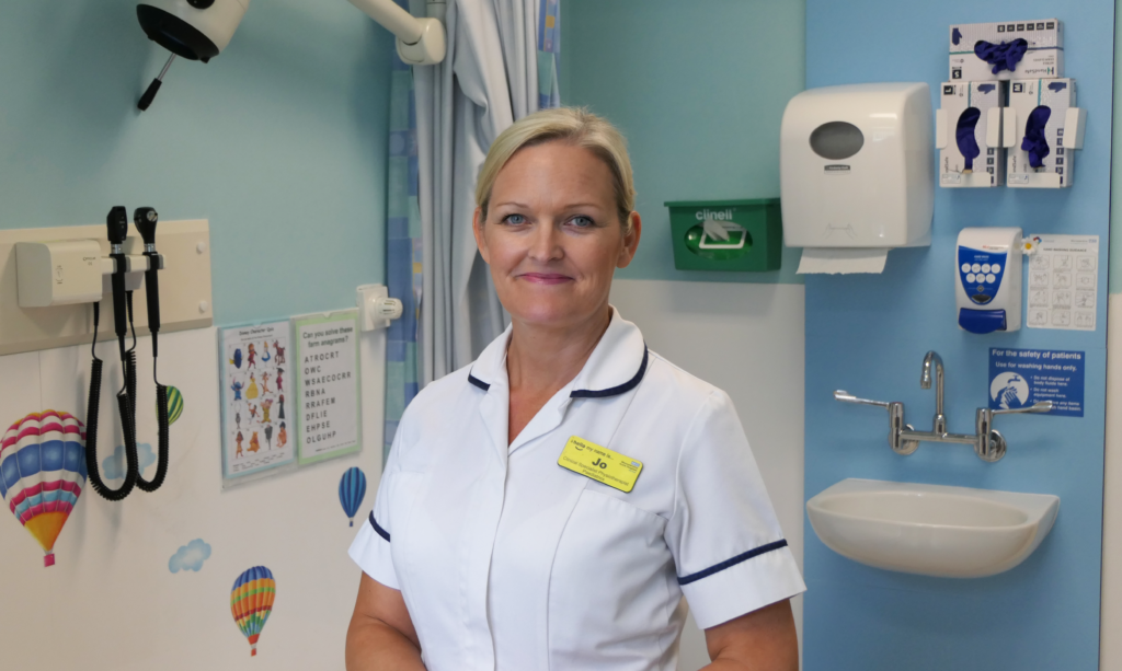 Healthcare professional in a white clinical uniform standing in a hospital examination room. The room includes a sink with soap and glove dispensers on the wall, medical instruments, and light blue walls decorated with small hot air balloon graphics.