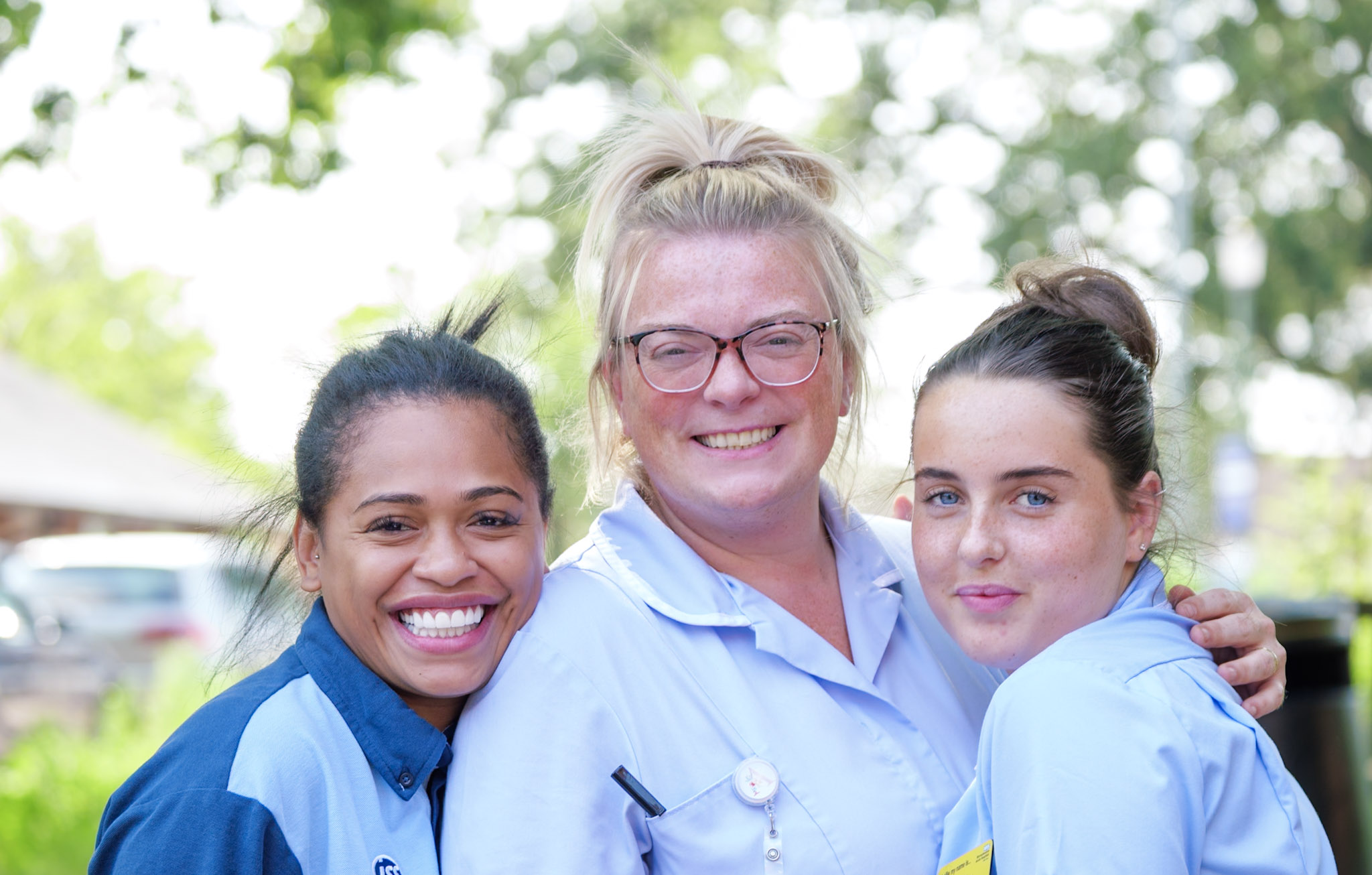 Three women smiling while working at WRH hospital, photographed outdoors with a softly blurred leafy background.