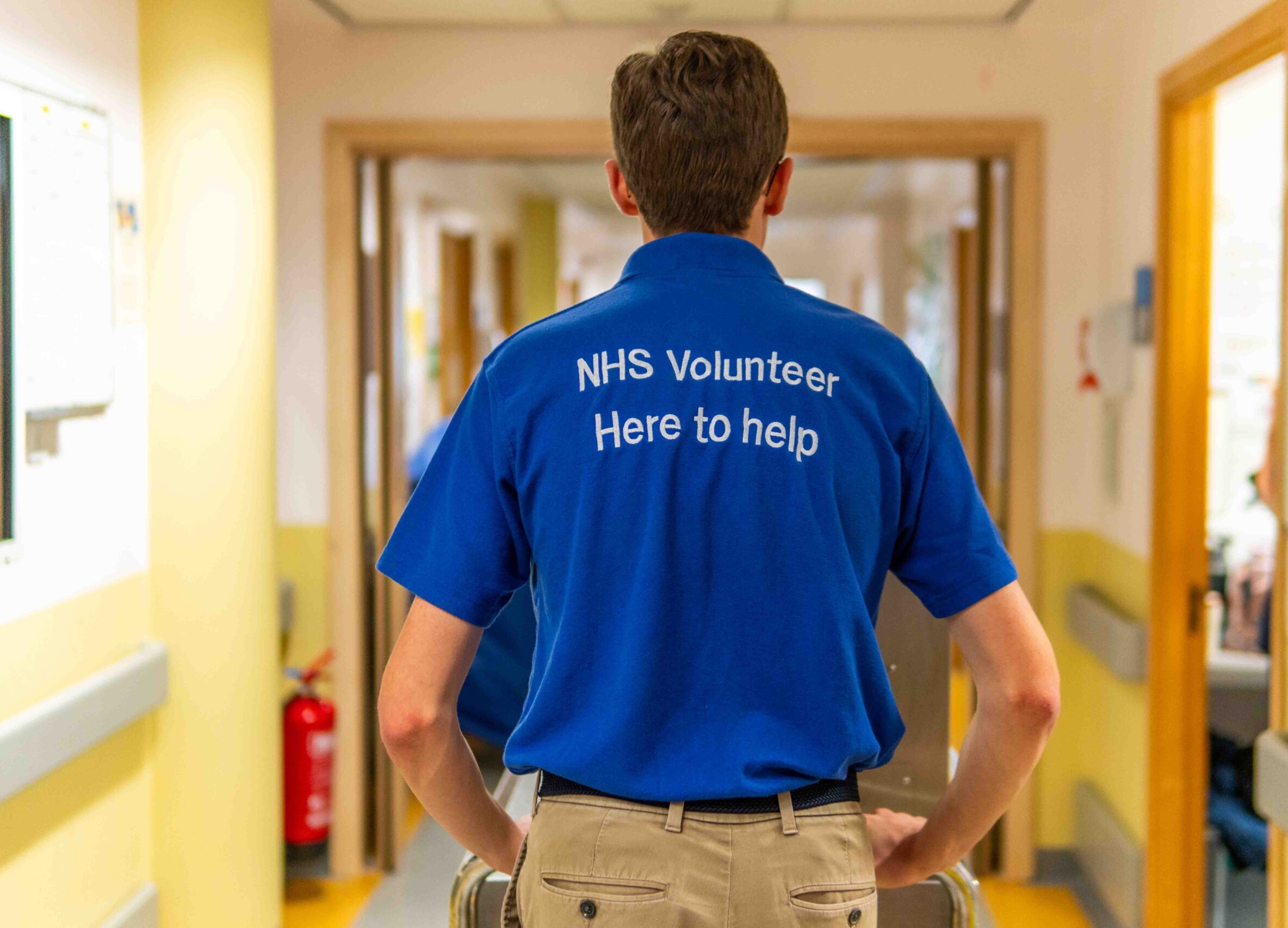 Photograph of a volunteer, focus on the writing on his polo short. NHS Volunteer. Here to help.