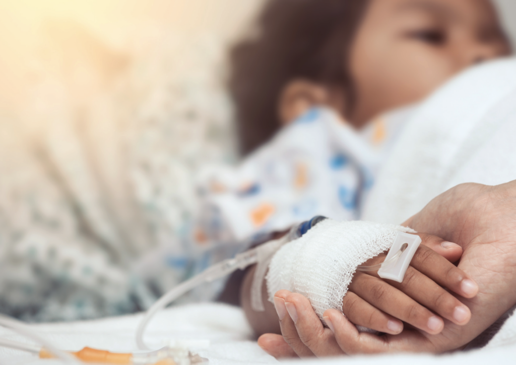 Close-up of a patient’s hand resting on a hospital bed, with an intravenous (IV) cannula secured by a bandage, while another hand gently holds it for support; the patient is wearing a patterned hospital gown in the background.