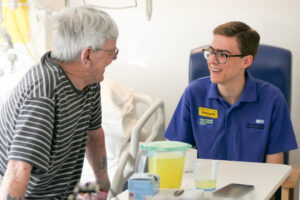 Hospital volunteer sitting by the side of a hospital bed talking and smiling with a patient