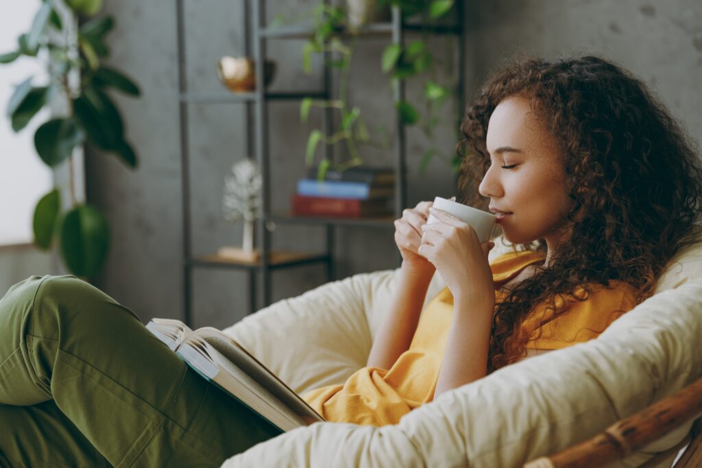 A relaxed woman drinking coffee and reading book with closed eyes sits in armchair at home.