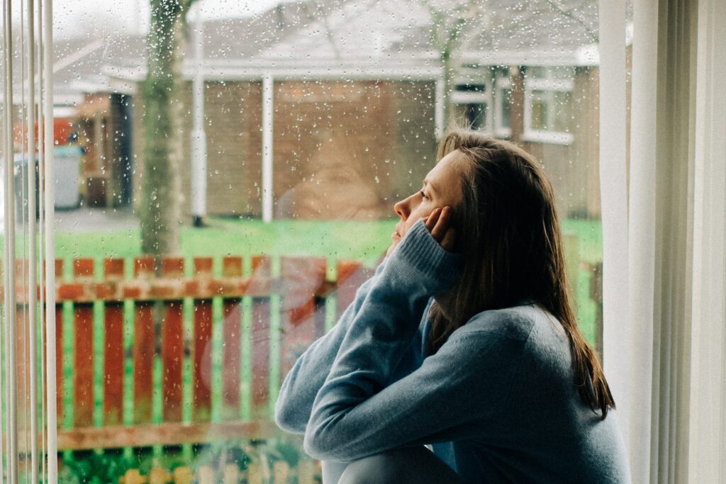Thoughtful sad woman sitting on a windowsill with a reflection on a window